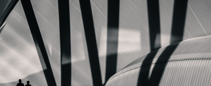 Two people silhouetted in front of a large curved metal structure with tall dark shadows behind them.