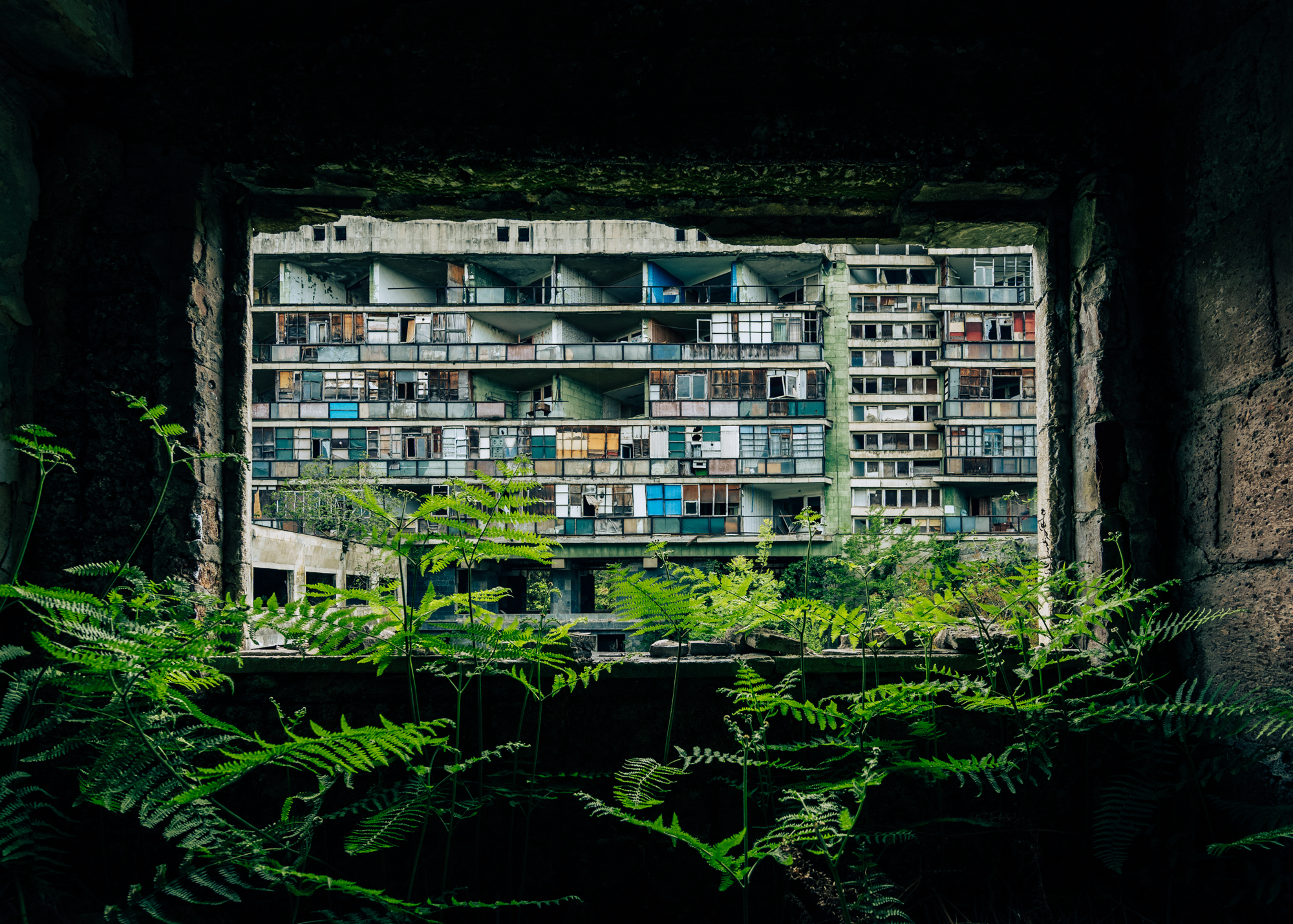 Abandoned multi-story apartment building seen through a dark cave, with green ferns in the foreground.